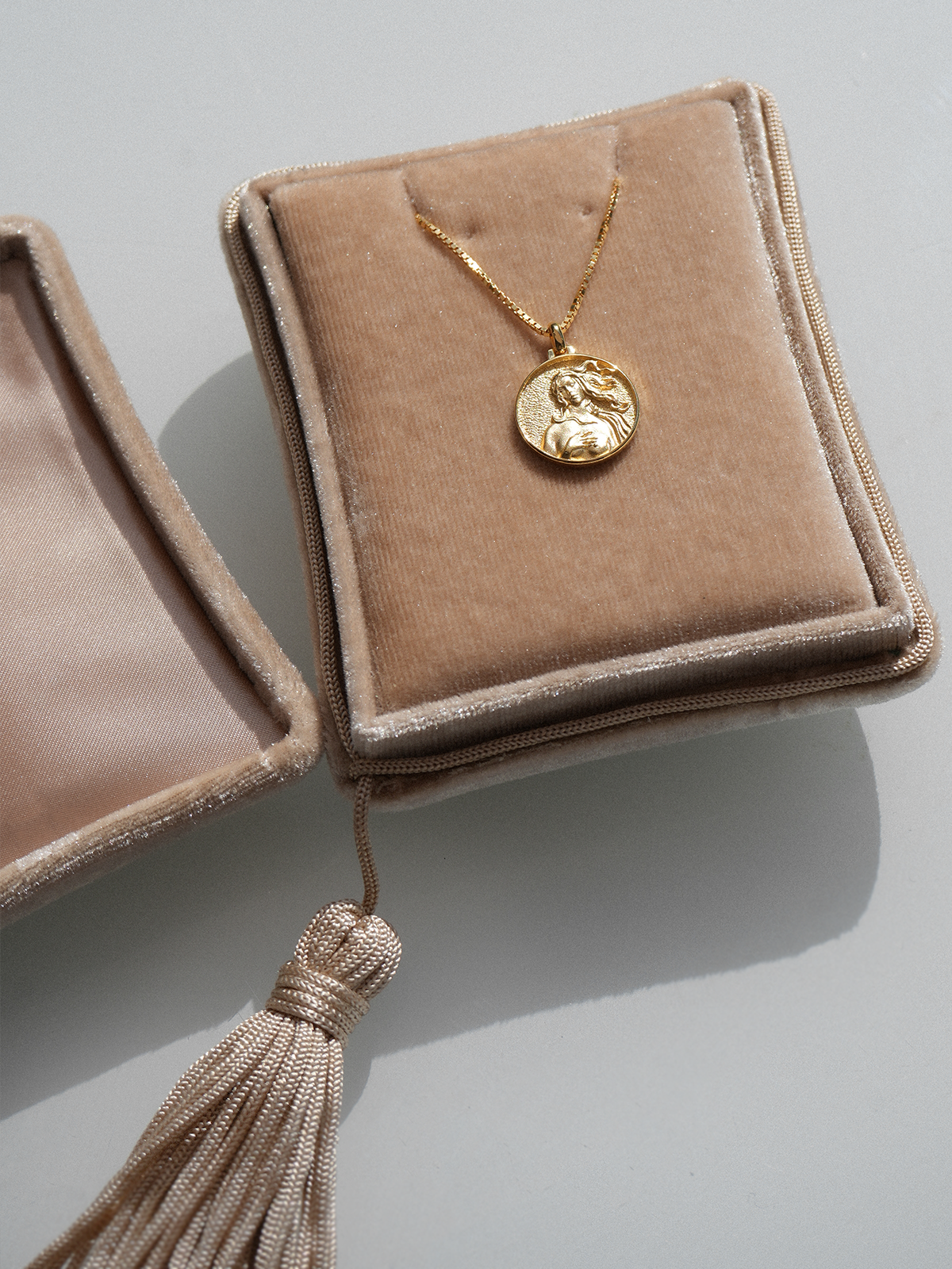 Beigefarbenes Goddess Jewelry Case aus Seidensamt mit dekorativer Quaste und goldener Kette auf einem Samtkissen.