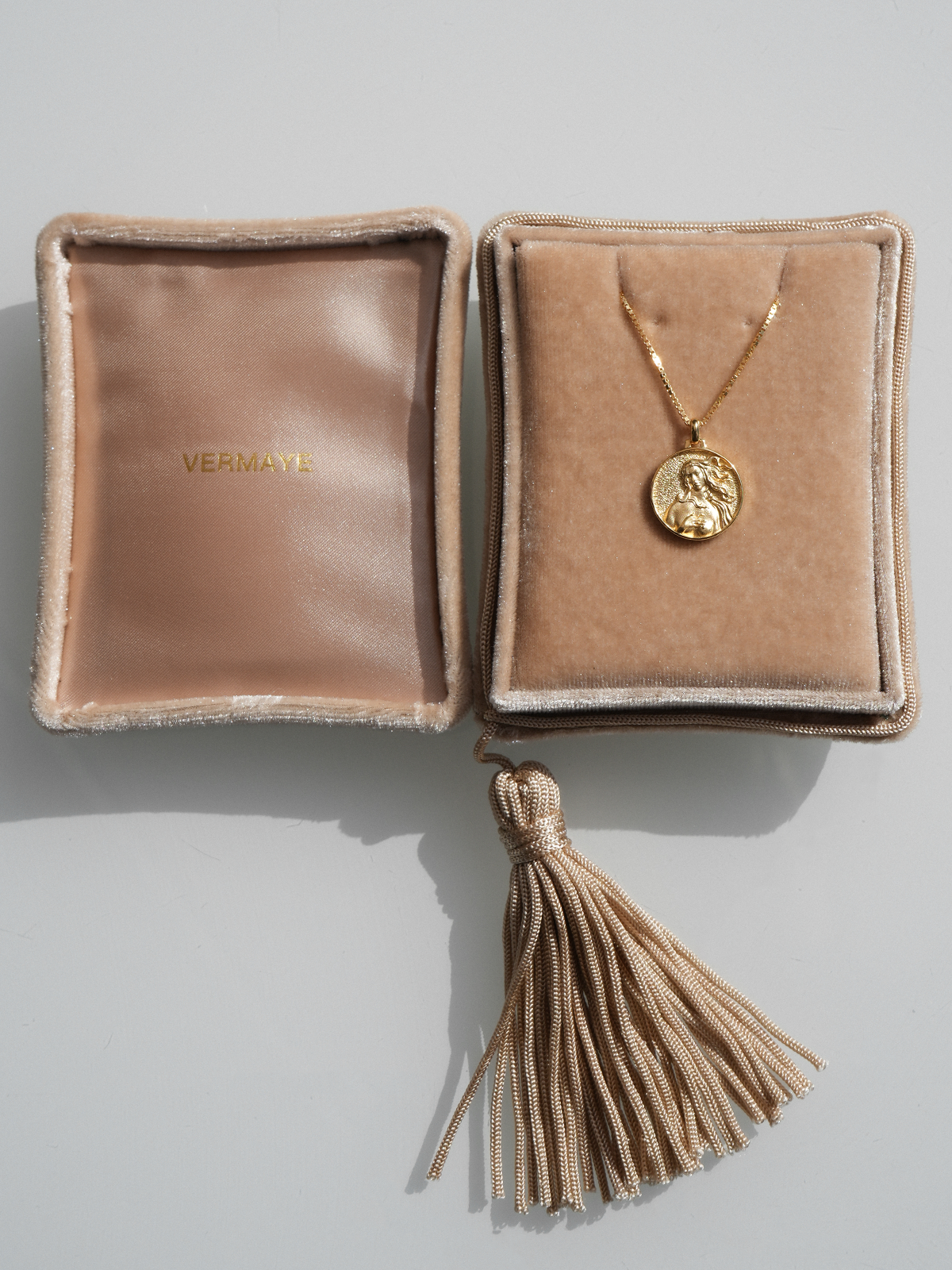 Beigefarbenes Goddess Jewelry Case aus Seidensamt mit dekorativer Quaste und goldener Kette auf einem Samtkissen.